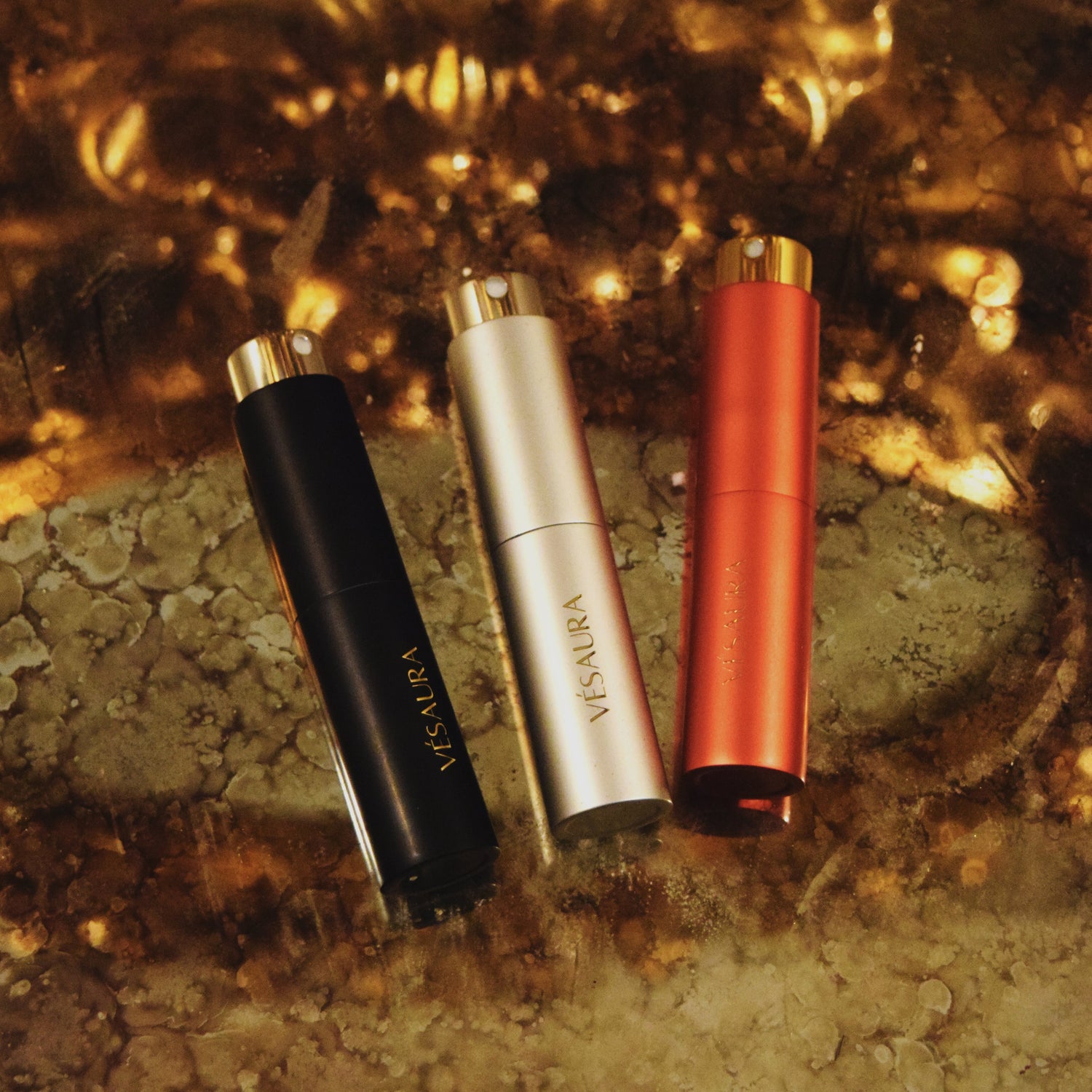 Three Vésaura perfume cases in silver, black, and red displayed against a gold luxury background, showcasing a bold and elegant trio of fragrances.
